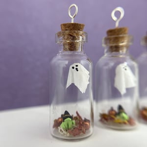 以下が含まれることがあります： コルク栓と銀色のワイヤーループが付いた小さなガラス瓶が3つ。各瓶には白い幽霊の置物と、魔女の帽子、カボチャ、葉など、ハロウィーンをテーマにした装飾が入っています。