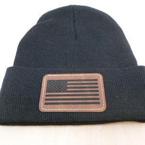 May include: A black knit beanie with a brown leather patch featuring the American flag. The patch is rectangular and stitched onto the front of the hat. The beanie is made of a soft, ribbed knit material.