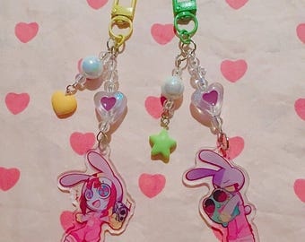 May include: Two acrylic keychains featuring cartoon rabbit characters in pink outfits. The left keychain has a yellow clasp, a yellow heart charm, and a clear heart bead. The right keychain has a green clasp, a green star charm, and a clear heart bead.