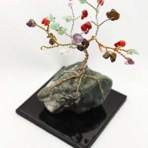 Könnte beinhalten: Ein drahtgewickelter Baum mit einer Vielzahl von bunten Edelsteinen, darunter Amethyst, Tigerauge und roter Jaspis, auf einem grauen Gesteinssockel. Der Baum steht auf einem schwarzen quadratischen Sockel.
