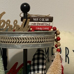 May include: A tiered tray decorated for Christmas with a gnome, a small pine tree, and a red glitter letter "M". The top tier has a black knob and three wooden signs stacked on top of each other that read "hot cocoa", "marshmallows", and "candy canes".