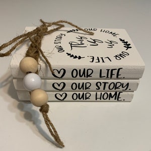 May include: A stack of four white painted wooden blocks with the words "This is our life. Our story. Our home."  The blocks are tied together with twine and decorated with wooden beads.