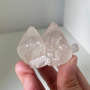 May include: A cluster of two clear, translucent, pink-hued crystals with a smaller, clear crystal attached to the side. The crystals have a rough, natural texture.