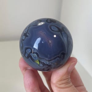 May include: A polished spherical stone featuring a deep blue-grey base color with intricate, darker circular and cloud-like patterns throughout its surface. The smooth, reflective orb displays natural mineral inclusions and subtle light reflections.