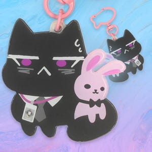 Puede incluir: Dos llaveros de acrílico con un personaje de gato negro de ojos morados, camisa blanca, corbata gris y una pequeña tarjeta de identificación, junto a un conejo rosa más pequeño con pajarita negra, ambos unidos a cierres de plástico rosa.