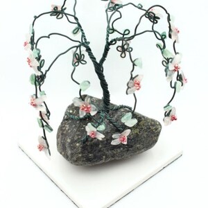 Könnte beinhalten: Eine Drahtskulptur eines Trauerweidens mit rosa und grünen Glasblumen. Der Baum steht auf einem grauen Steinsockel.