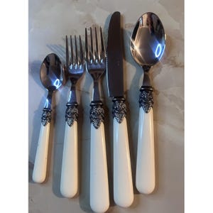 May include: A five-piece flatware set consisting of a small spoon, two forks, a dinner knife, and a large spoon. Each piece features a polished stainless steel head, an ornate dark metal collar with leaf-like detailing, and a smooth off-white handle.