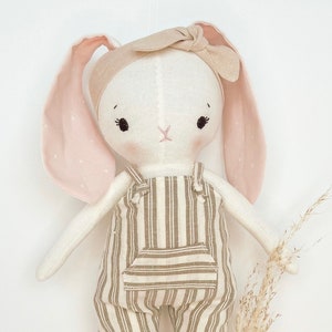 May include: A white stuffed bunny wearing a brown and white striped overall and a light brown headband. The bunny has pink ears and black eyes.