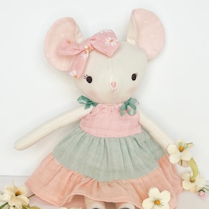 May include: A white plush mouse doll wearing a pink dress with green and pink layers. The doll has a pink bow with white flowers in its hair and is sitting on a white surface with white flowers.