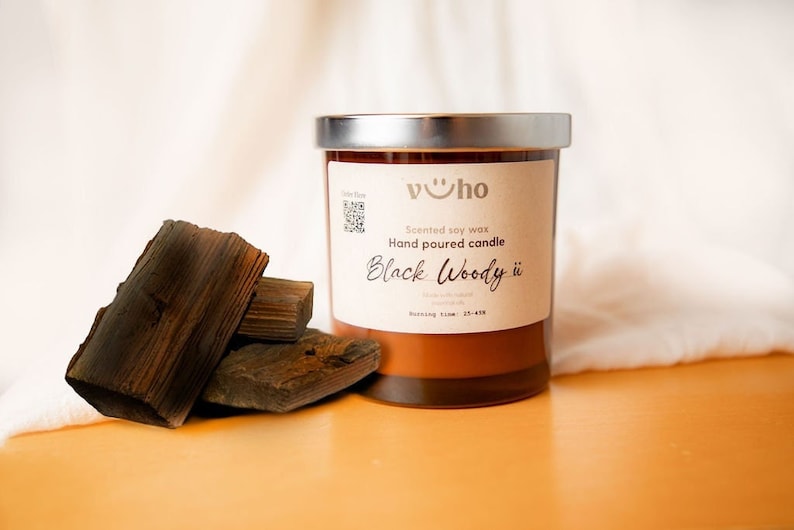 Puede incluir: Un tarro de vela de vidrio marr&oacute;n con una tapa plateada. La etiqueta dice "Cera de soja perfumada Vela vertida a mano Black Woody ii Tiempo de combusti&oacute;n: 25-45h". Un mont&oacute;n de trozos de madera marr&oacute;n oscuro se encuentran en primer plano.