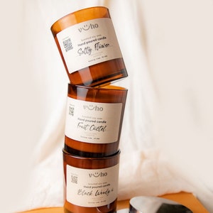 Puede incluir: Tres tarros de velas de vidrio marr&oacute;n con tapas apiladas uno encima del otro. Los tarros est&aacute;n etiquetados con el texto "vho" y las fragancias "Sally Flower", "Fruit C&uuml;ctel" y "Black Woody".