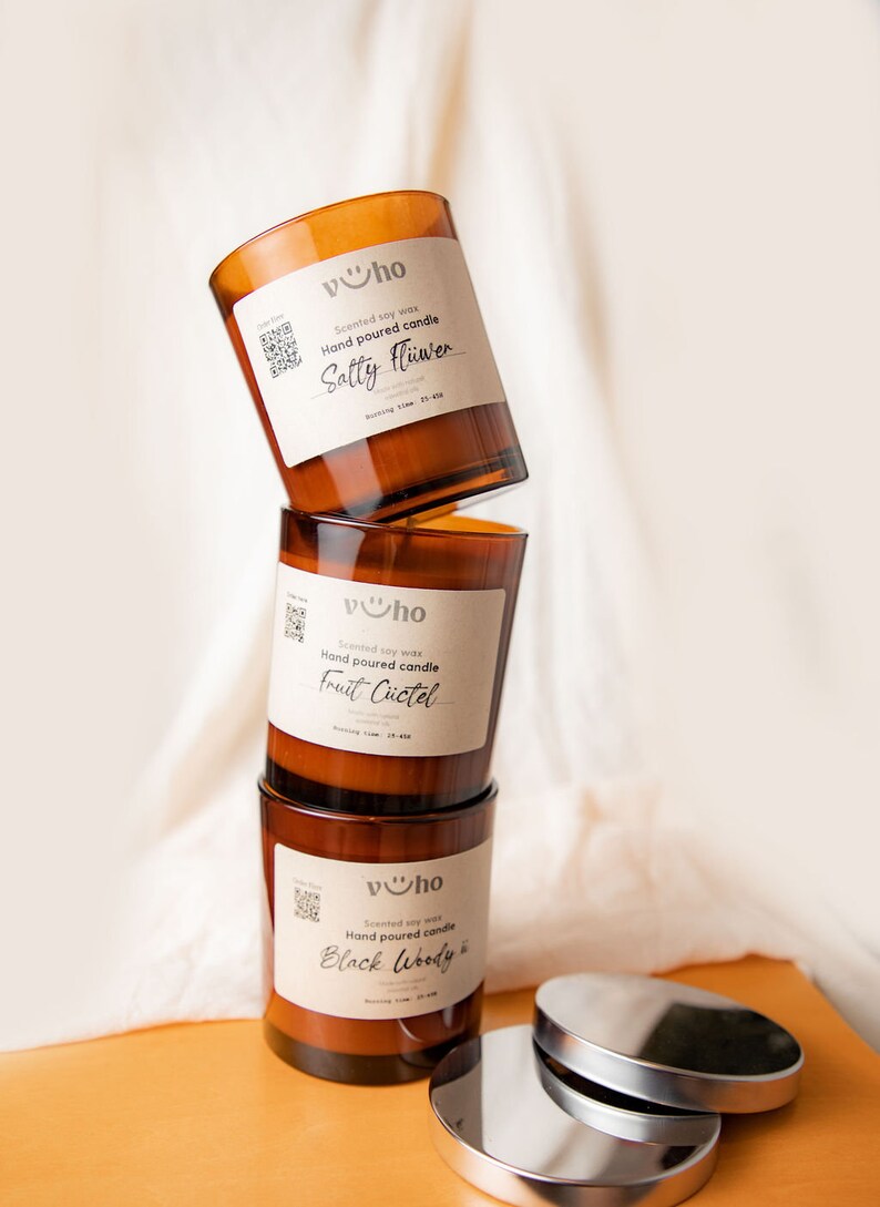 Puede incluir: Tres tarros de velas de vidrio marr&oacute;n con tapas apiladas uno encima del otro. Los tarros est&aacute;n etiquetados con el texto "vho" y las fragancias "Sally Flower", "Fruit C&uuml;ctel" y "Black Woody".