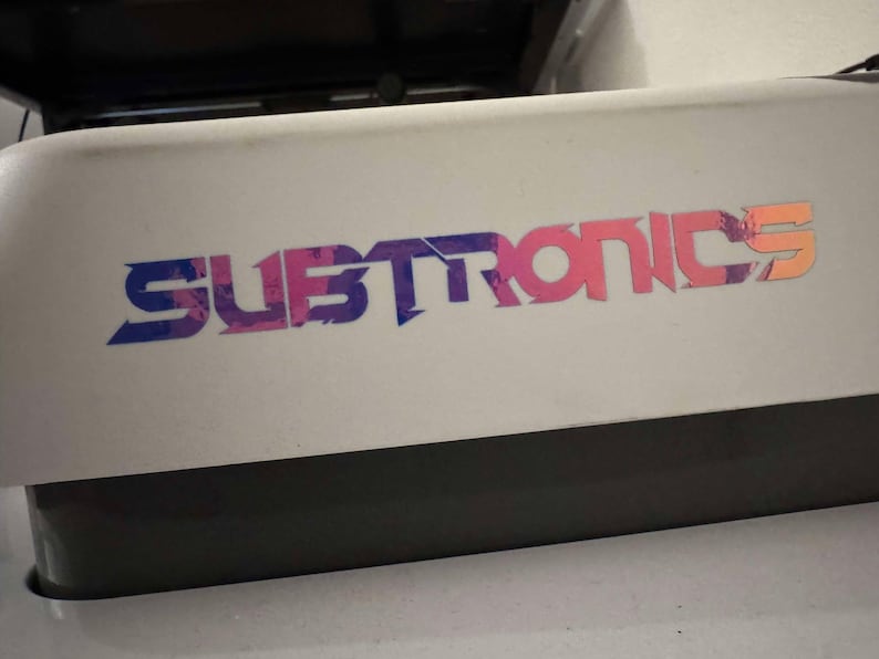 Puede incluir: Una superficie blanca con un logotipo colorido que dice "SUBTRONICS" en una fuente estilizada con colores rosa, azul y naranja.