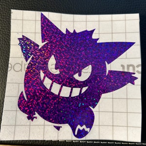 May include: A purple and blue glitter vinyl sticker of a cartoon ghost with a mischievous grin. The sticker is on a white gridded background.