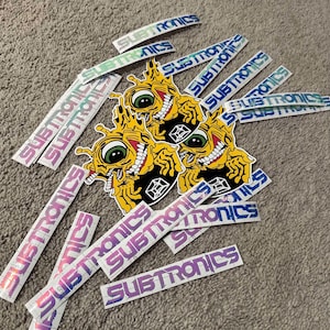Puede incluir: Una colecci&oacute;n de pegatinas con un personaje de dibujos animados con un ojo grande y una sonrisa de dientes. Las pegatinas est&aacute;n impresas con la palabra "Subtronics" en una variedad de colores y estilos.