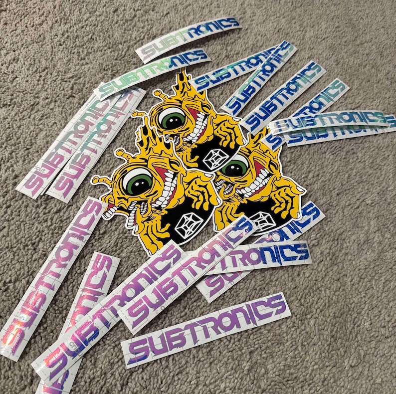 Puede incluir: Una colecci&oacute;n de pegatinas con un personaje de dibujos animados con un ojo grande y una sonrisa de dientes. Las pegatinas est&aacute;n impresas con la palabra "Subtronics" en una variedad de colores y estilos.