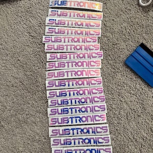 Puede incluir: Una pila de 15 pegatinas blancas con la palabra "SUBTRONICS" impresa en un degradado de rosa y azul.