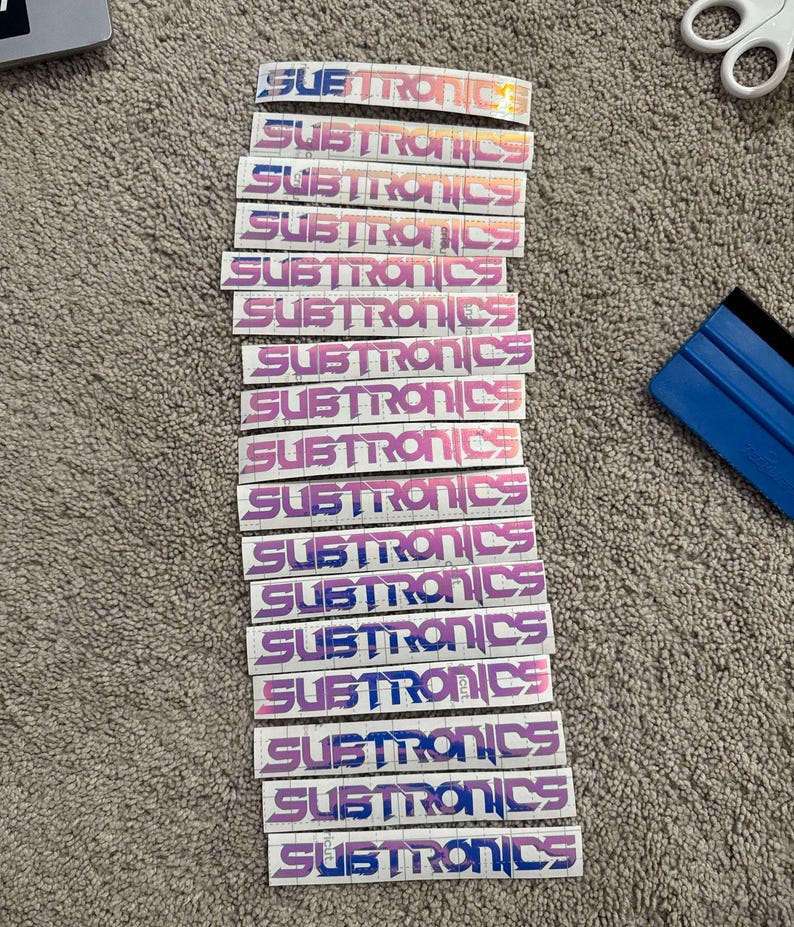 Puede incluir: Una pila de 15 pegatinas blancas con la palabra "SUBTRONICS" impresa en un degradado de rosa y azul.