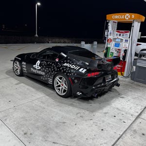 May include: A black sports car with white and red rims is parked at a gas station at night. The car has a large rear spoiler and is covered in white graphics and the PlayStation logo. The gas station has a Circle K sign.