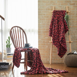 May include: A red and black leopard print blanket draped over a wooden chair and a ladder. A black teacup and saucer sit on a book. A cactus in a gray pot is on the left. The background is a brick wall and a window.