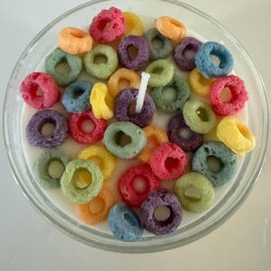 May include: A clear glass bowl filled with a white liquid and colorful, ring-shaped cereal pieces. A white candle is centered in the bowl. The cereal pieces are in various colors, including red, purple, green, and yellow.