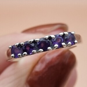 Può includere: Un anello a fascia d'argento con cinque pietre preziose viola incastonate in fila.