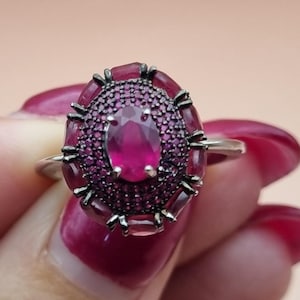 May include: A silver ring featuring a large oval ruby gemstone, surrounded by smaller ruby stones. The ring has a detailed design with intricate silverwork.