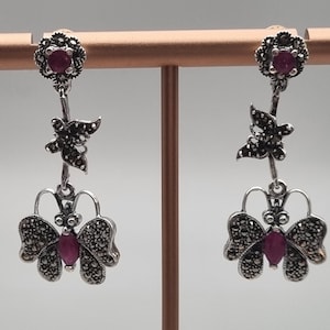 May include: A pair of silver earrings with a dangling butterfly design. Each earring features a red gemstone in the centre of the butterfly and a smaller butterfly charm above it.