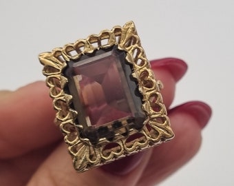 May include: A gold ring featuring a large, rectangular, purple gemstone set within an ornate, decorative frame.