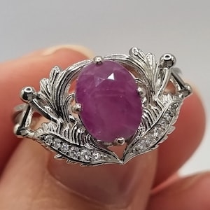 Op de afbeelding: Een zilveren ring met een grote ovale roze edelsteen, omringd door kleinere heldere edelstenen en een delicaat bloemmotief.