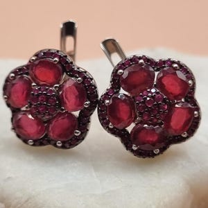May include: A pair of silver earrings with a flower design. Each earring features five red ruby stones set in a silver flower shape.