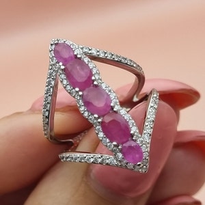 May include: A silver ring with a row of five pink oval gemstones set in a silver arrow design.