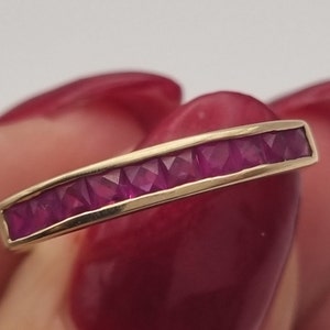 May include: A gold band ring with a row of small, square-shaped ruby stones.