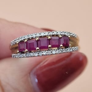 May include: A gold ring featuring a central row of five square-cut deep red rubies held by prongs, flanked by two parallel bands encrusted with small, clear round-cut diamonds. The design showcases a split-shank structure with a polished metal finish.