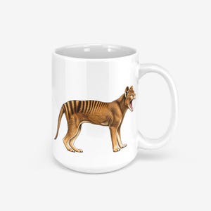 Könnte beinhalten: Ein weißer Keramikbecher mit einer detaillierten Illustration eines Beutelwolfs, auch bekannt als Tasmanischer Tiger. Das Tier ist im Profil mit weit geöffnetem Maul dargestellt und zeigt sein beiges Fell sowie markante dunkelbraune Streifen.