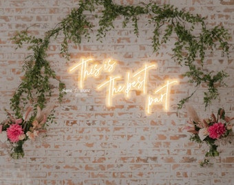 May include: A white brick wall with greenery hanging from the top. A neon sign with the text "This is the best part" is mounted on the wall. The sign is lit up with a warm yellow glow. The text "Save the Date" is written below the sign.