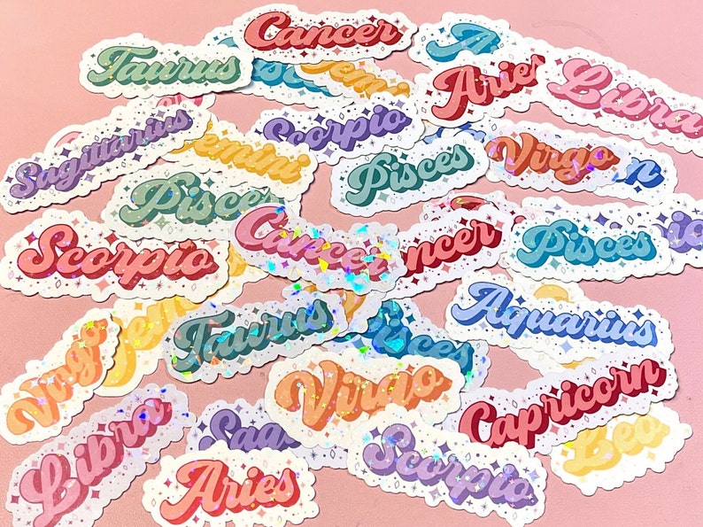 May include: A collection of colorful, glittery stickers featuring the astrological signs of the zodiac. The stickers are arranged in a random pattern on a pink background. The signs include: Taurus, Cancer, Aries, Libra, Sagittarius, Gemini, Scorpio, Pisces, Virgo, Aquarius, Capricorn, Leo.