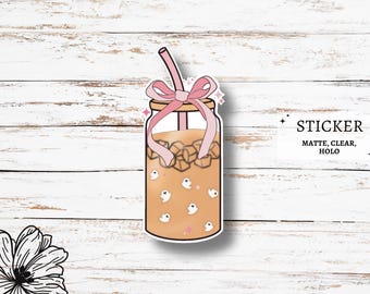 May include: A sticker of a mason jar filled with iced coffee and ice cubes. The jar has a pink ribbon tied around the lid and a pink straw sticking out. There are small white ghosts floating in the coffee. The sticker says "STICKER MATTE, CLEAR, HOLO" at the bottom.