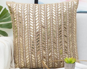 Puede incluir: Una almohada de sofá beige y dorada con un patrón de chevron. La almohada está en un sofá blanco con una manta blanca y una bandeja dorada con una pequeña planta en maceta.