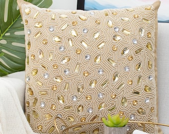 May include: A gold and cream throw pillow with a beaded design. The pillow is sitting on a couch with a throw blanket and a wooden tray with a plant and glasses.