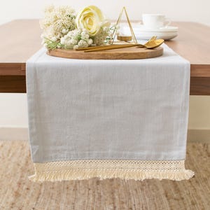 May include: A light blue rectangular table runner featuring a cream-colored crocheted lace border with fringe trim. The fabric has a smooth, matte texture and hangs over the edge of a wooden table, providing a neutral base for table decor.