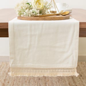 May include: A rectangular off-white table runner featuring a decorative crocheted lace border and a fringe edge. The fabric is smooth and solid in color, draped over the edge of a wooden table, providing a neutral base for table settings.