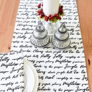 May include: A white table runner with black text that reads "Believe, Claus, Cheer, Jingle Bell, Fa La La, Holly, Jolly, Naughty or Nice, Mistletoe, Snowman, Sugar, Plummy, Tis the Season, Jingle Bells, Noel".  Two silver candle holders with a white candle in the center are on the table runner.