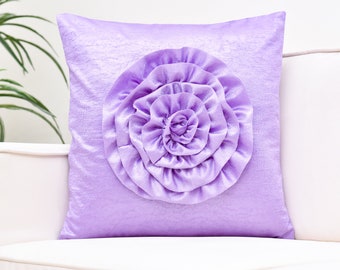 May include: A square lavender pillow with a large, ruffled flower design in the center. The pillow has a soft, textured appearance and is placed on a white sofa. The flower is a lighter shade of lavender.