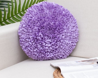 May include: A round, purple, ruffled throw pillow on a white couch. The pillow is made of soft fabric and has a delicate, floral design.
