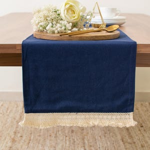 May include: A solid navy blue table runner featuring a cream-colored lace trim with fringe along the bottom edge. The runner is draped over a wooden table, providing a flat surface for a round wooden board, white flowers, and gold cutlery.