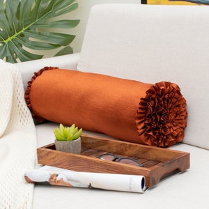 May include: A rust-colored cylindrical pillow with a ruffled edge sits on a white couch. A small wooden tray with a succulent plant and glasses sits on the couch in front of the pillow.