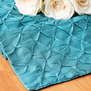 May include: Teal blue table runner with a quilted, gathered design. The runner is made of a soft, silky fabric and has a subtle sheen.