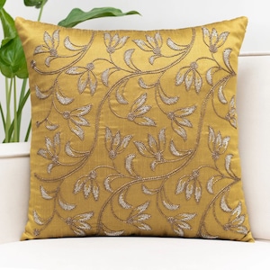 May include: A mustard yellow throw pillow with a floral pattern embroidered in silver thread.