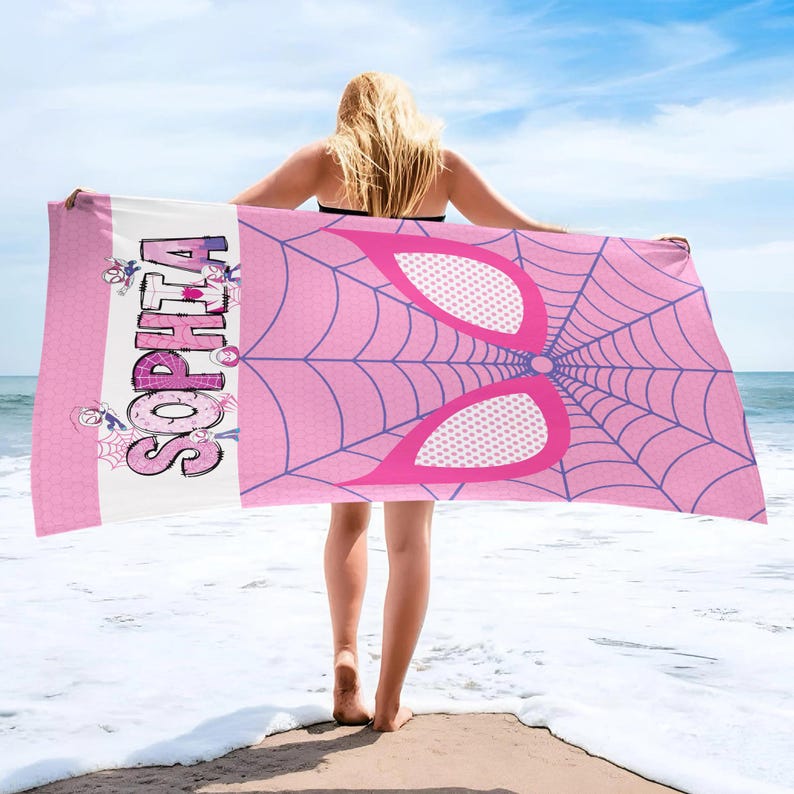 May include: A pink beach towel featuring a spider web design and the name "SOPHIA" in a purple and white font. The towel has a pink and white border with cartoon spider graphics. The background shows the ocean and sky.